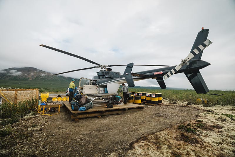 A helicopter parked on a makeshift wooden platform surrounded by fuel drums, with gear and personnel preparing for departure in a remote area.