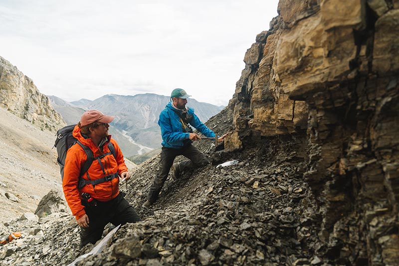 Two geologists collect rock samples from a cliff in a mountainous area, with rugged peaks in the background.