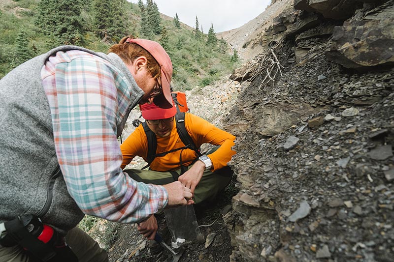 Dr Ross Anderson wearing outdoor gear examine a rocky cliff face, one using a small tool to collect rock samples.