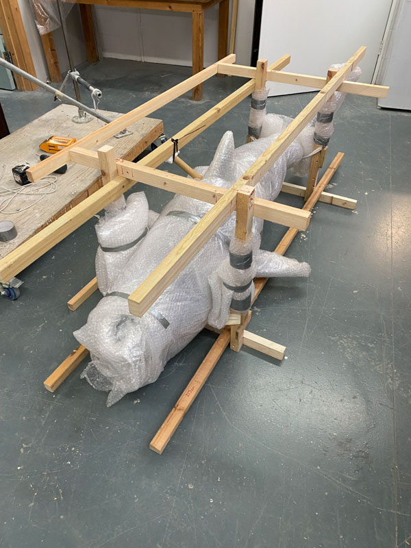 A set of timber beams set a right angles, acting as a frame to carry a model sitting between them. The model is not visible directly as it is wrapped in plastic packing material including bubble wrap.