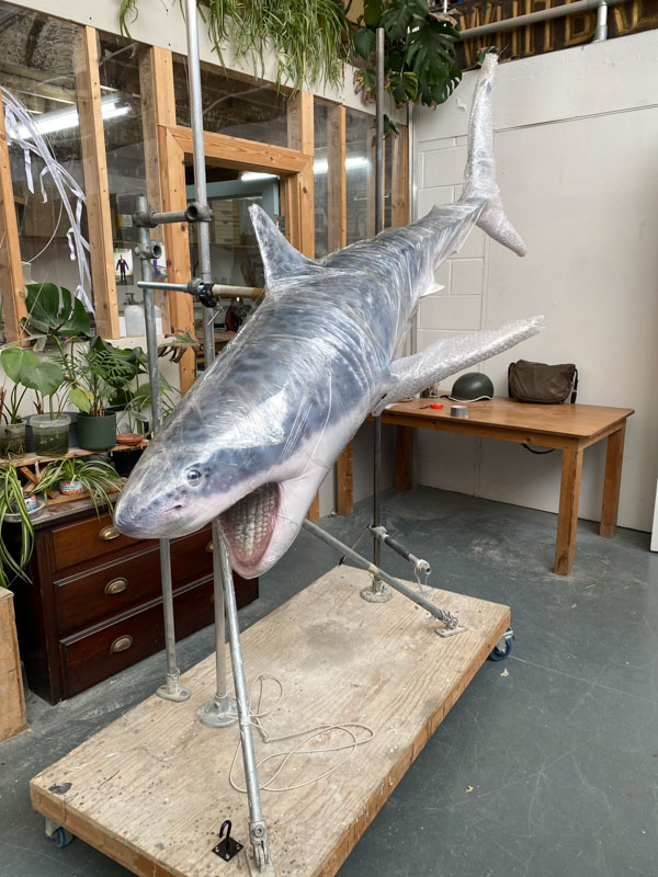 A fibreglass shark model mounted on a stand horizontally, wrapped in a clear plastic. In the background is workshop.